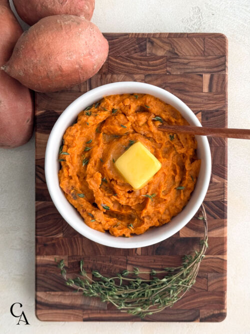 Mashed sweet potatoes with fresh thyme and a square of butter, styled with whole sweet potatoes on a wooden cutting board.