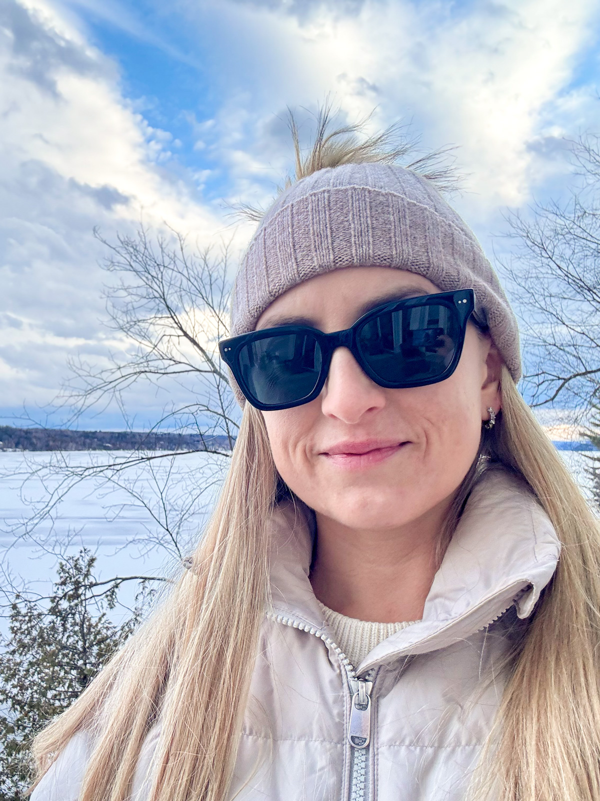 A blonde woman wearing sunglasses and a hat, standing in front of a frozen lake in winter.