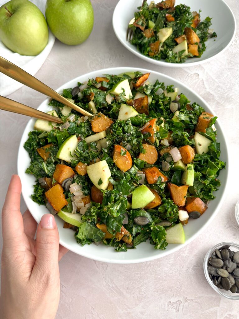 Overhead view of a kale salad with roasted acorn squash, green apple chunks, sautéed shallots, pumpkin seeds, and hemp seeds in a white bowl with gold serving utensils.