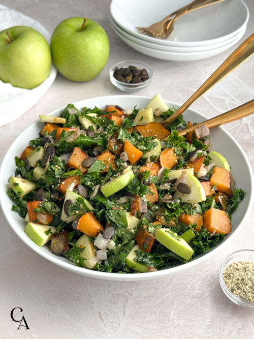 Styled bowl of salad with kale, squash, green apple pieces and seeds, photographed on a neutral surface with apples and bowls in the background.
