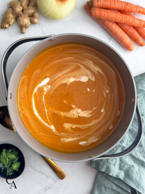 Dutch oven filled with blended carrot and ginger soup, showing a creamy orange texture before garnishing, with fresh carrots nearby.
