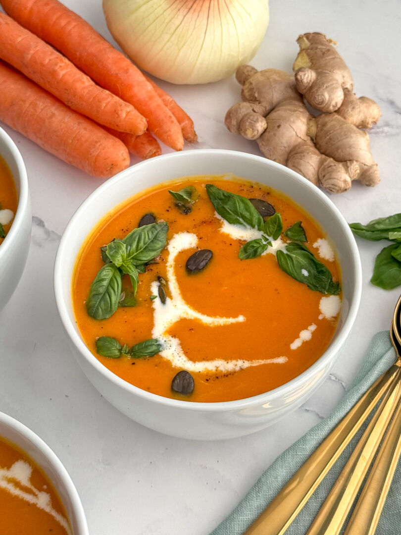 Bowl of carrot ginger soup garnished with fresh basil, pumpkin seeds, and a swirl of almond milk, with whole carrots, onion, and fresh ginger in the background.