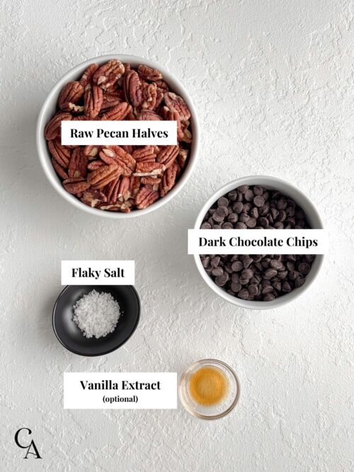 Bowls of raw pecan halves, dark chocolate chips, flaky salt, and vanilla extract arranged on a light surface.