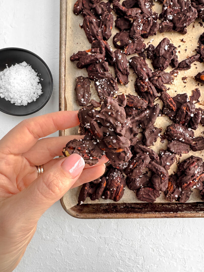 Hand holding a cluster of dark chocolate covered pecans sprinkled with flaky sea salt over a parchment-lined baking sheet.