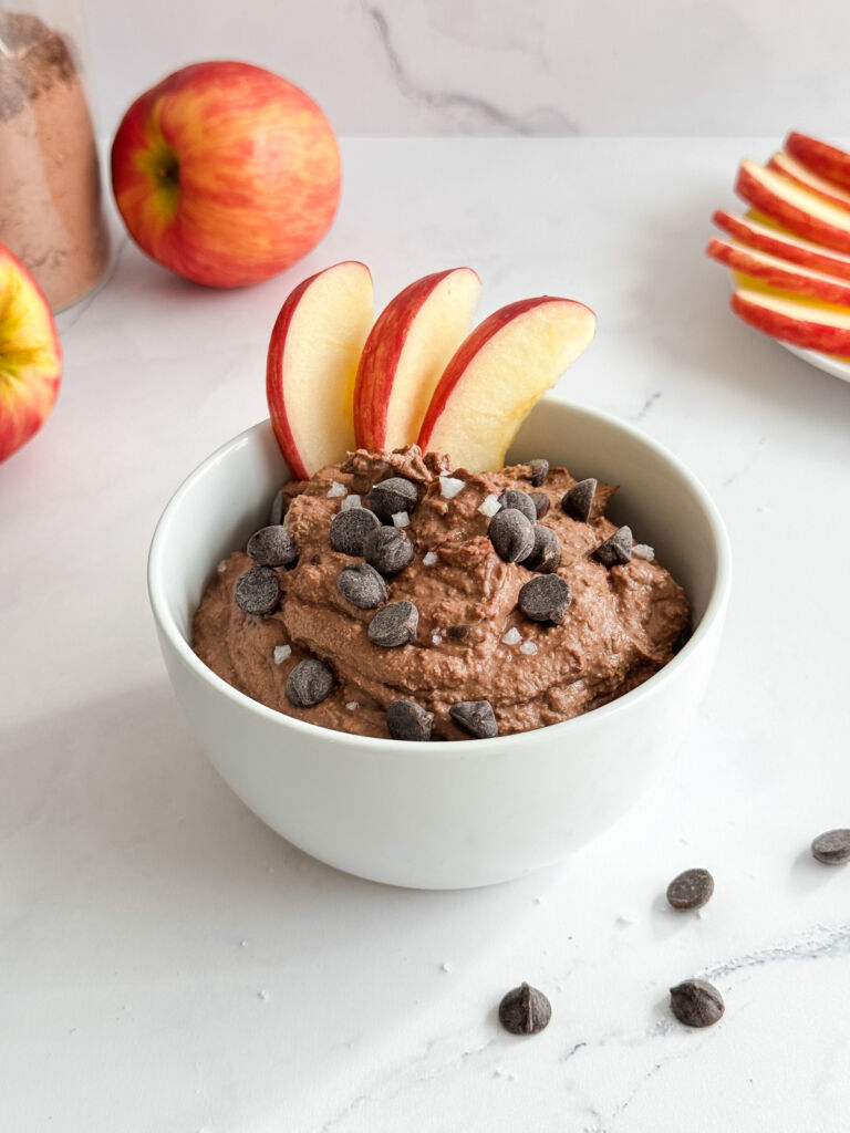 A small bowl of chocolate hummus topped with chocolate chips, flaky sea salt, and red apple slices.