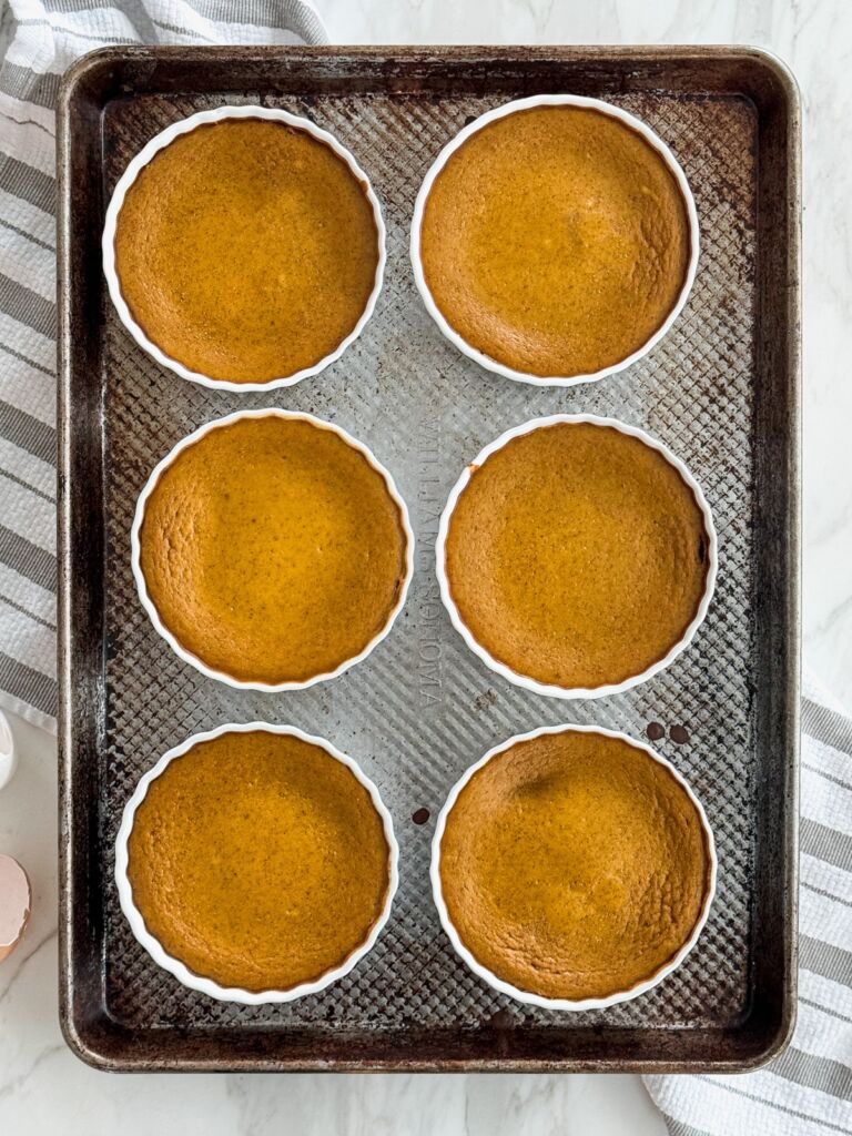 A rimmed baking sheet holding six pumpkin custards, on top of a white and grey striped towel.