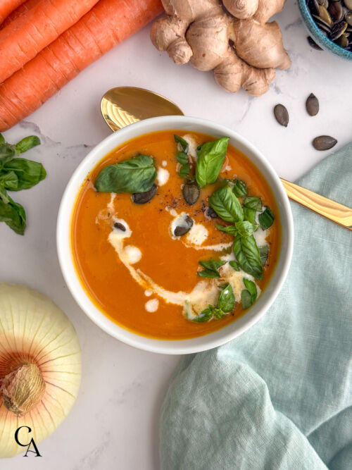 Healthy carrot soup in a white bowl topped with basil and pumpkin seeds, served with a gold spoon and fresh vegetables on a linen napkin.