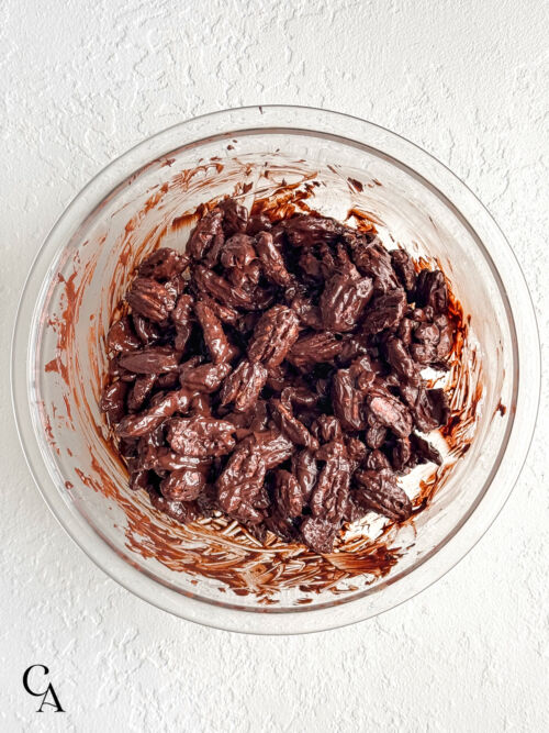 Pecans fully coated in melted dark chocolate inside a glass mixing bowl.