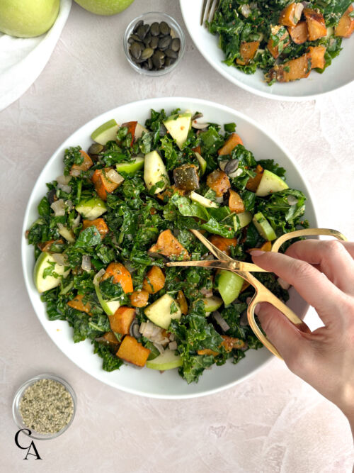 Top view of kale apple salad with roasted acorn squash and sautéed shallots being tossed with gold tongs in a white serving bowl.