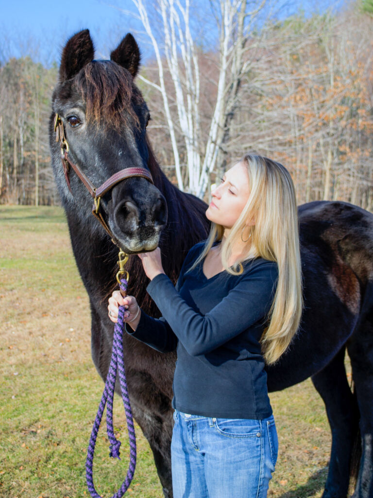 A young, blonde woman stands with her dark bay horse, holding a purple leadline attached to her leather halter.