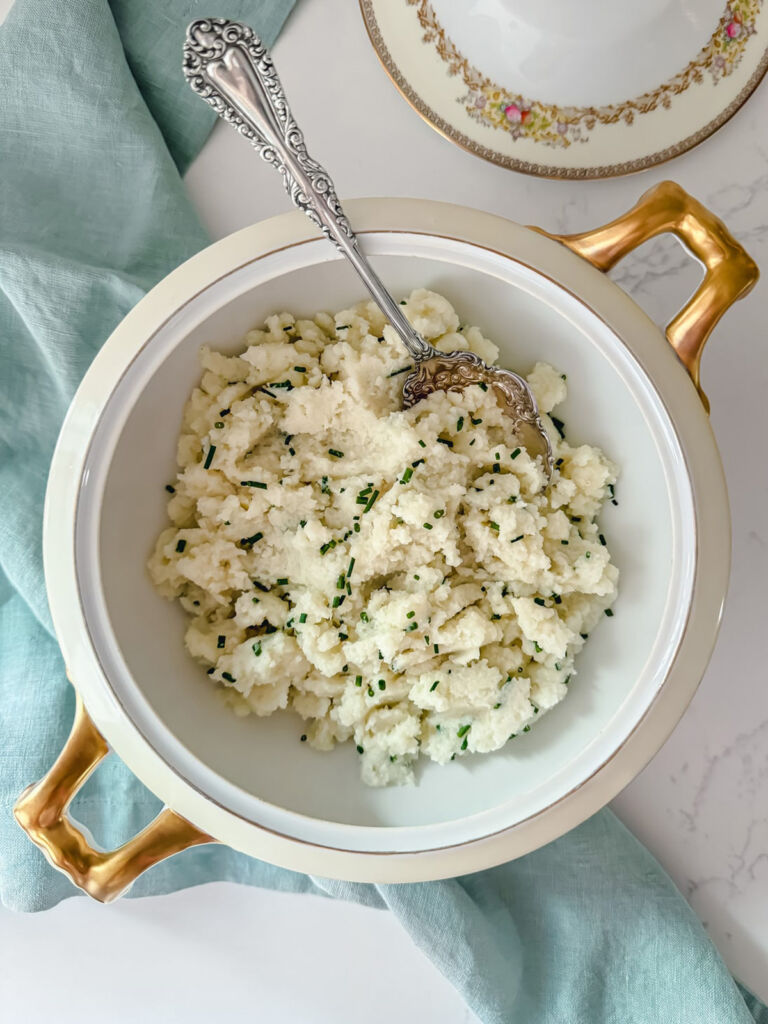 A china serving bowl full of mashed cauliflower sprinkled with finely chopped chives, and a silver spoon.