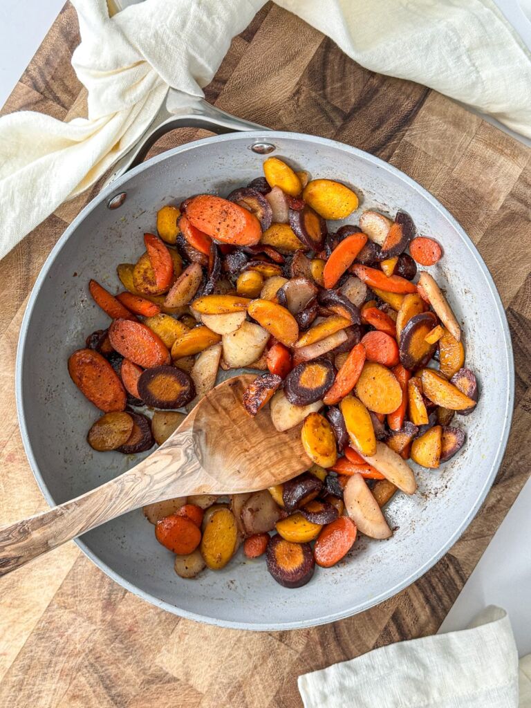 A large pan of sautéed rainbow carrots sitting on a wooden board, with a wooden spoon resting inside.