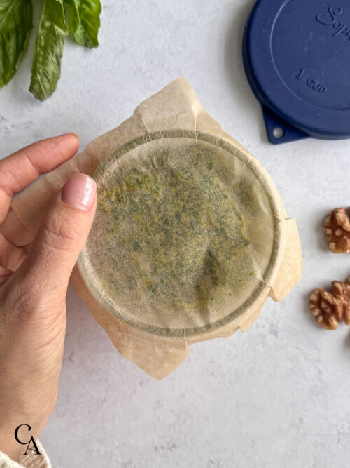 A woman's hand holding a piece of parchment paper over a glass container of pesto.