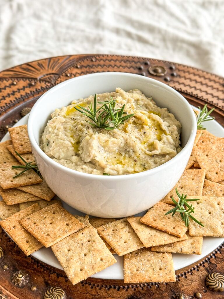 A white bowl of white bean dip, centered on a plate of crackers, topped with fresh rosemary and a drizzle of olive oil.