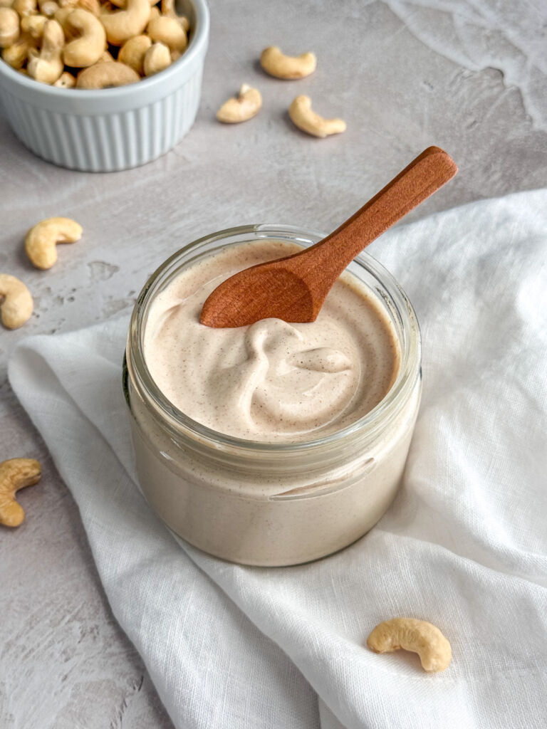 A glass jar of homemade cashew cream with flecks of cinnamon throughout, and a small wooden spoon resting inside.