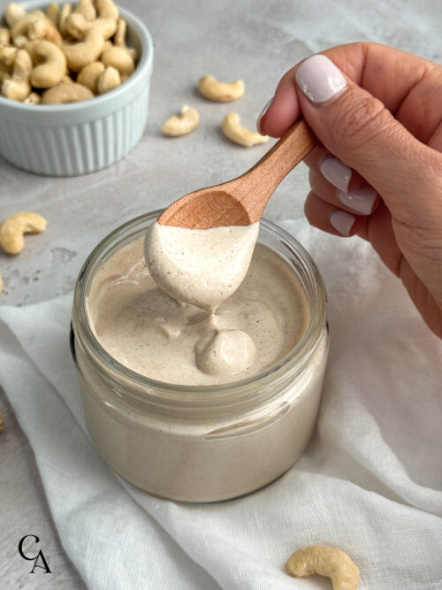 A manicured hand lifts a small wooden spoonful of sweet cashew cream from a glass jar.