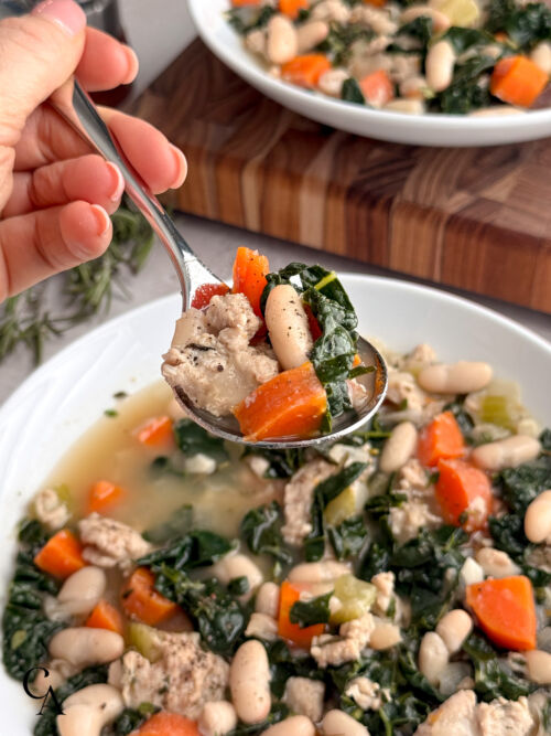 A silver soup spoon lifts chicken, cannellini beans, kale, and carrots from a bowl of soup.