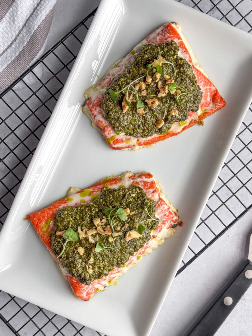 Baked pesto salmon fillets on a white plate topped with chunky basil pesto and chopped nuts.