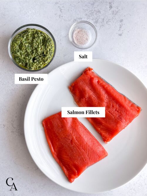 Raw salmon fillets on a white plate, with bowls of basil pesto and salt next to it.