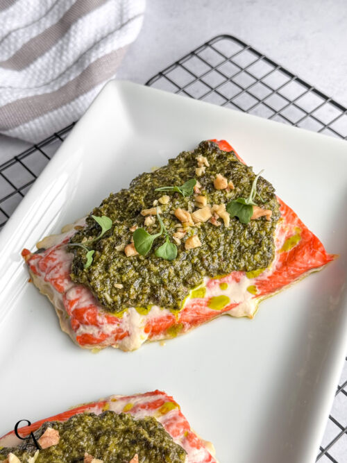 Close-up of baked salmon topped with pesto, chopped walnuts, and fresh greens on a white plate.