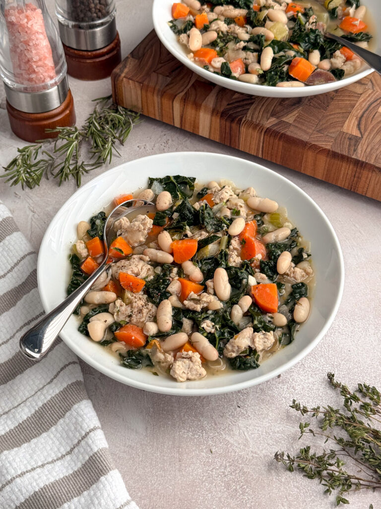 Bowl of hearty white bean, kale, and chicken soup with carrots and herbs.