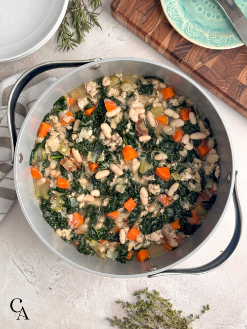 A large pot filled with a chunky soup of ground chicken, beans, and vegetables, next to a wooden cutting board.