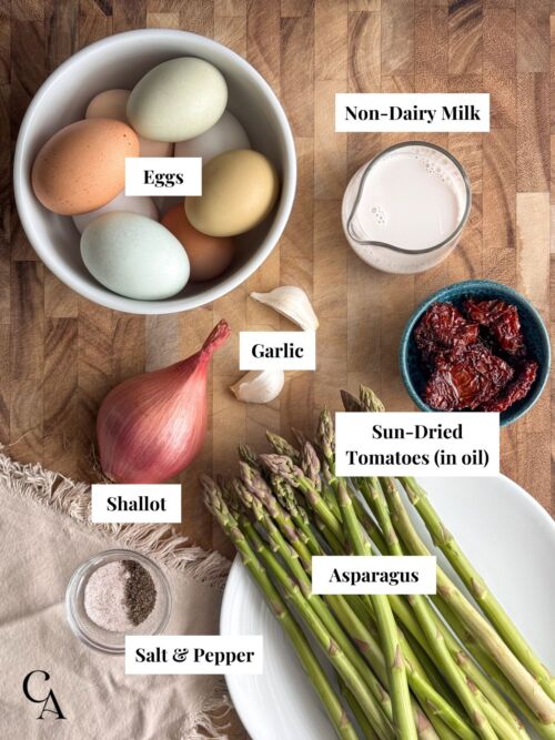 Ingredients in bowls, including eggs, asparagus, garlic, shallot, almond milk, and sun-dried tomatoes on a wooden surface.