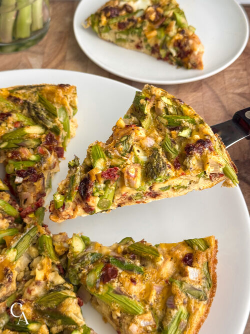 A slice of frittata with asparagus on a serving knife, with additional slices on a plate below.