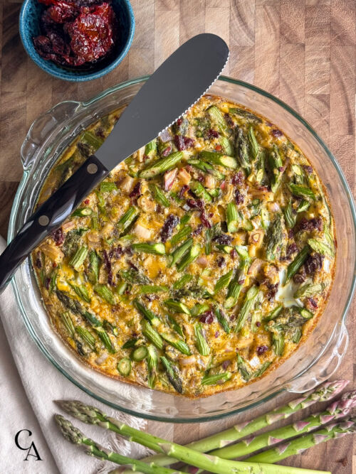 Freshly baked asparagus frittata in a glass pie dish, with a knife ready for slicing and a bowl of sun-dried tomatoes on the side.