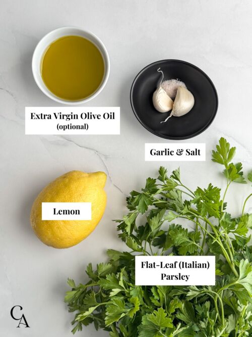 Bowls of ingredients on a marble surface, including olive oil, garlic, lemon, and a bunch of flat-leaf parsley.