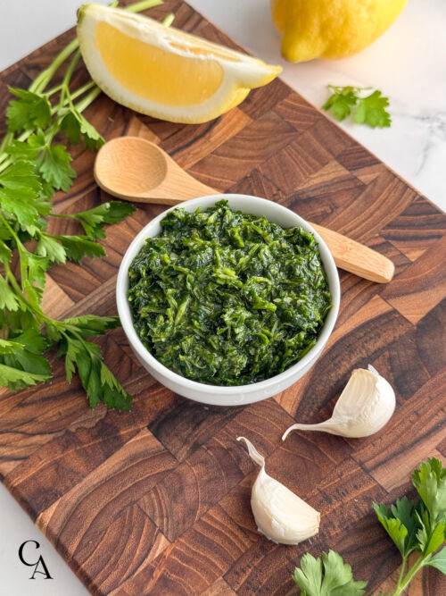 A small bowl of gremolata sauce next to a bamboo condiment spoon, garlic, parsely and wedges of lemon.