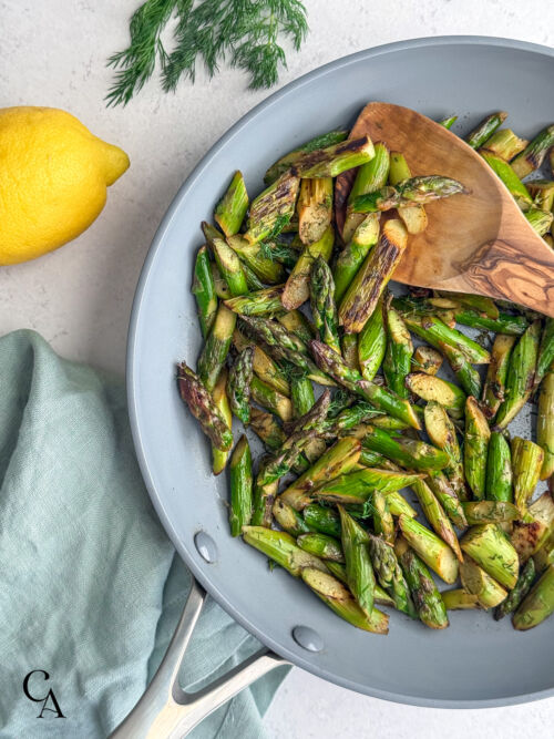 A close-up of a skillet of freshly sautéed asparagus next to a lemon and a bunch of dill.
