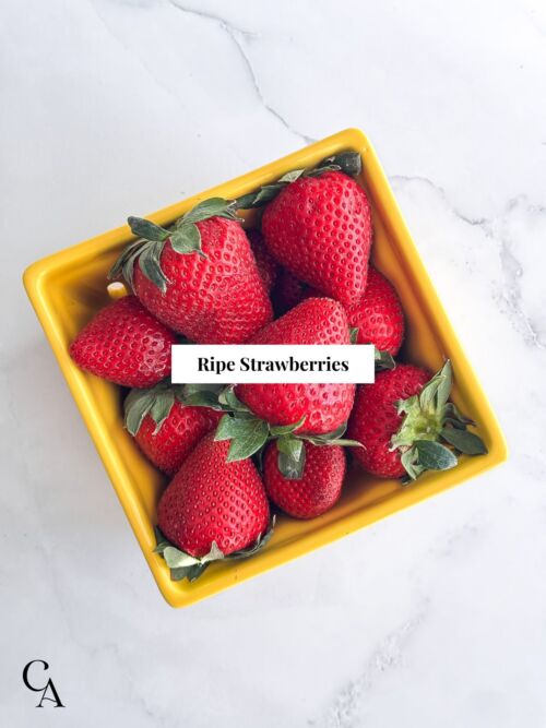 Overlay of a square yellow container of fresh strawberries on a marble countertop.