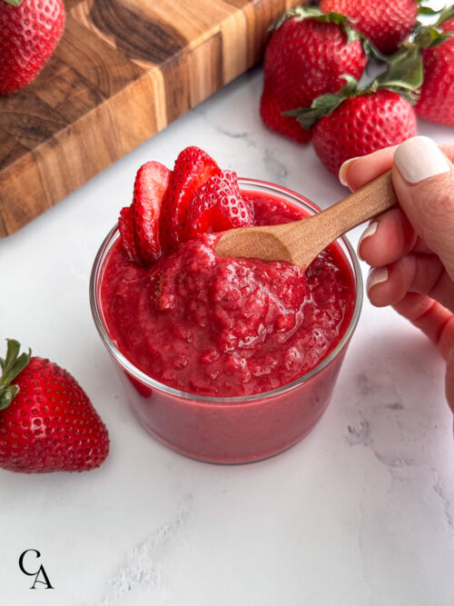 A woman's hand is stirring fresh strawberry sauce on a marble surface.