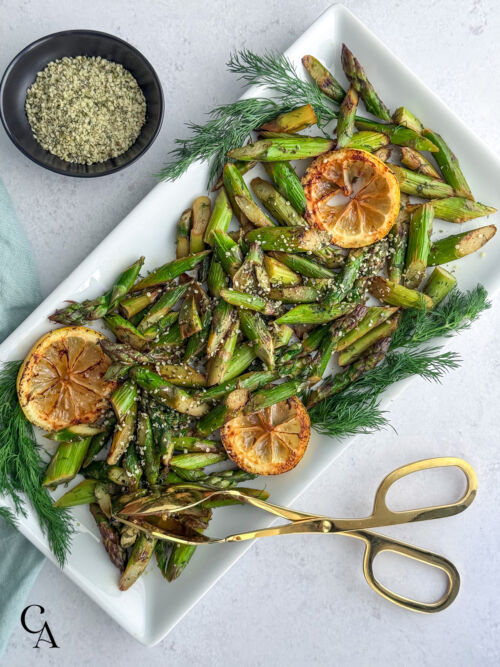 A platter of cut and cooked asparagus garnished with fresh dill, sautéed lemon slices and hemp seeds on top.