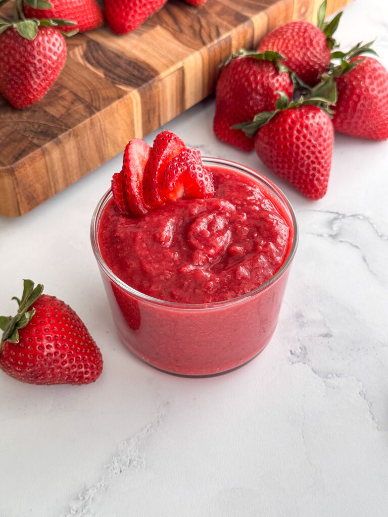 Fresh strawberry purée in a glass bowl, surrounded by whole strawberries with green tops.