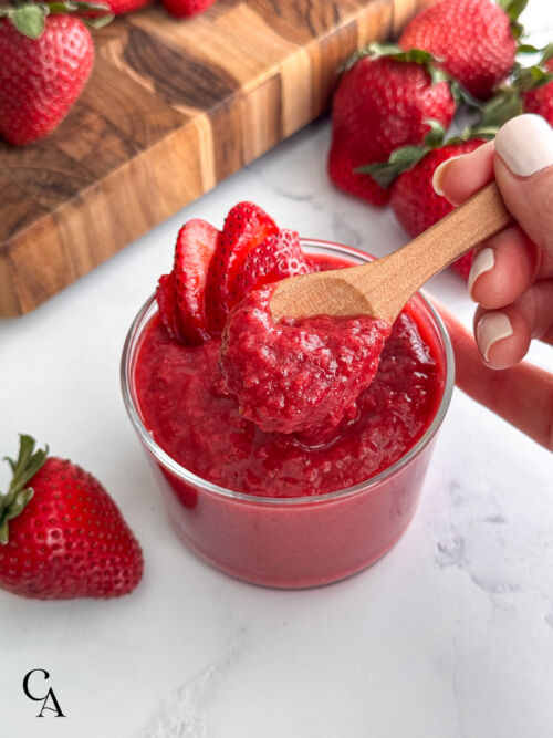 A hand lifting a spoonful of strawberry-purée from a jar.