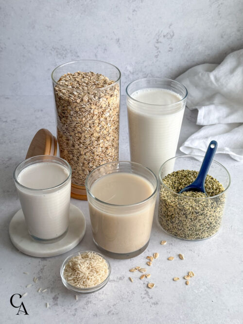 A canister of oatmeal and hemp seeds arranged with three glasses of non-dairy milk.