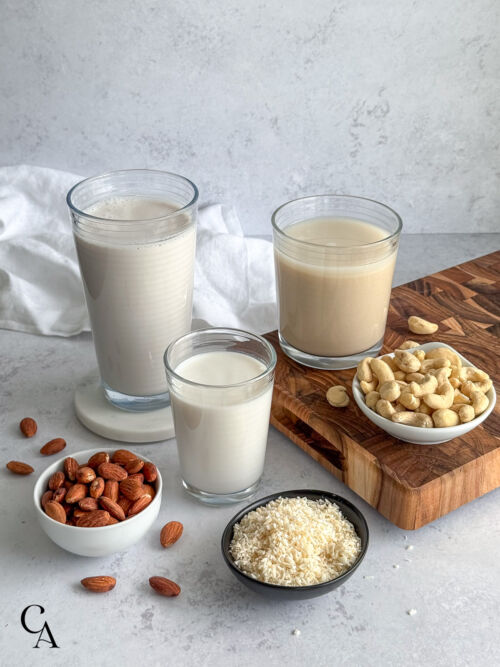 Glasses of almond milk, cashew milk and coconut milk on a light gray surface, with a wooden board on the side.