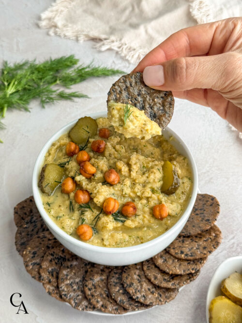 A woman's hand dipping a cracker into a bowl of pickle hummus topped with fresh dill.