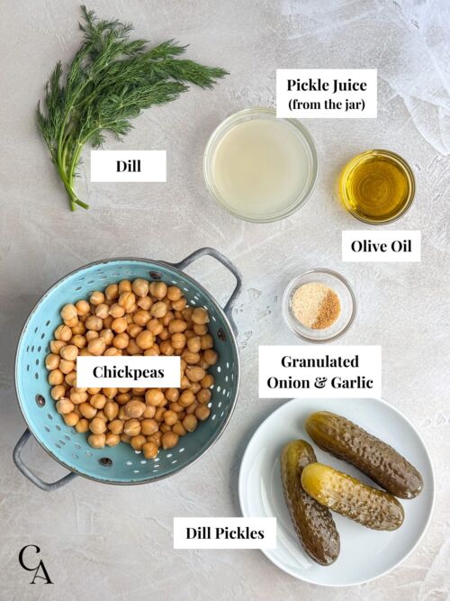 Chickpeas in a colander, next to a plate of pickles, a bunch of fresh dill, and small bowls of pickle juice, olive oil, and spices.