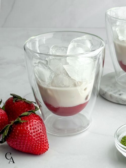 A glass with strawberry purée in the bottom, a layer of almond milk, and ice cubes next to fresh berries.