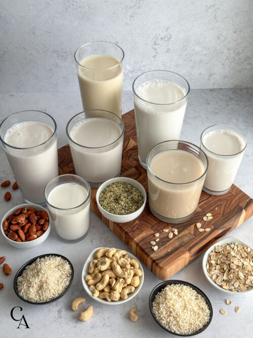 Glasses of different vegan milk options on a wood board, with bowls of nuts and grains in front.