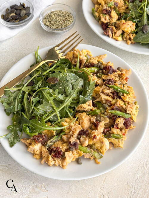 A plate of scrambled eggs with crab and veggies, plated next to a small breakfast salad topped with seeds.