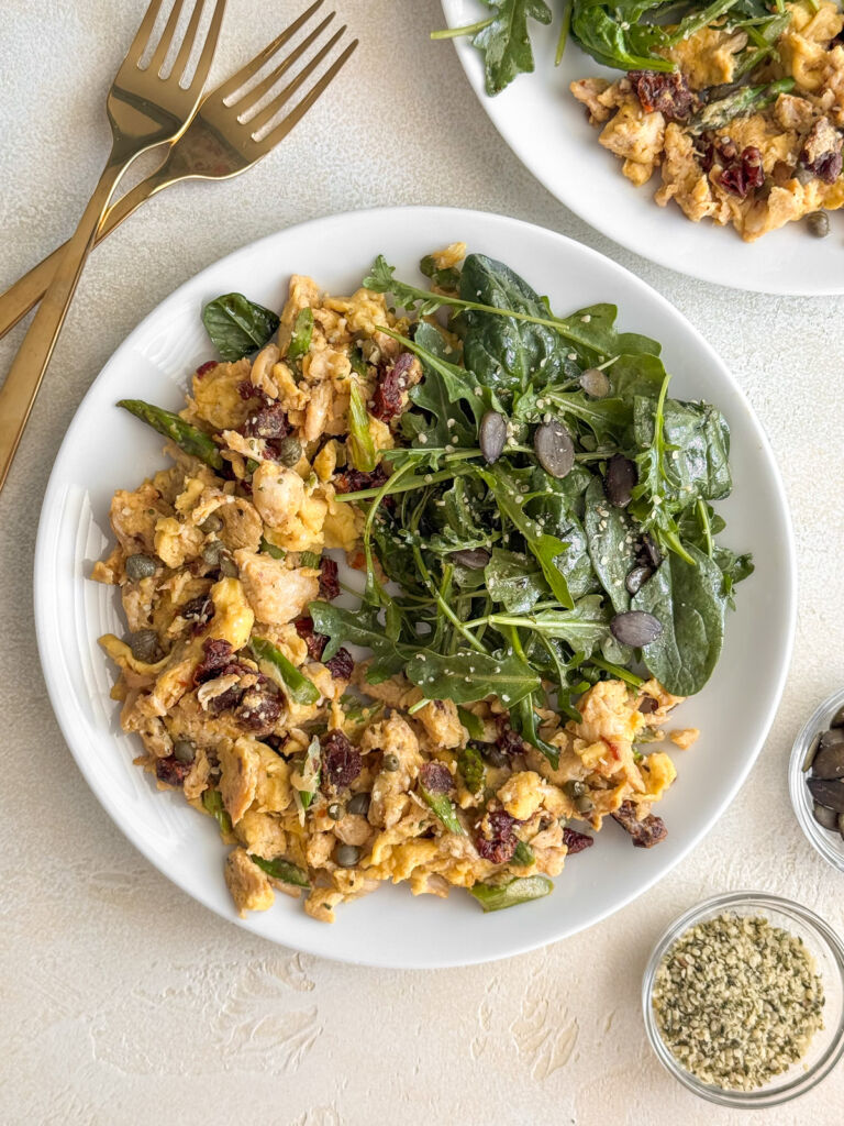 Flat-lay of a plate of breakfast eggs with crab meat and fresh veggies, next to a small leafy green salad, with gold forks and bowls of seeds on the side.
