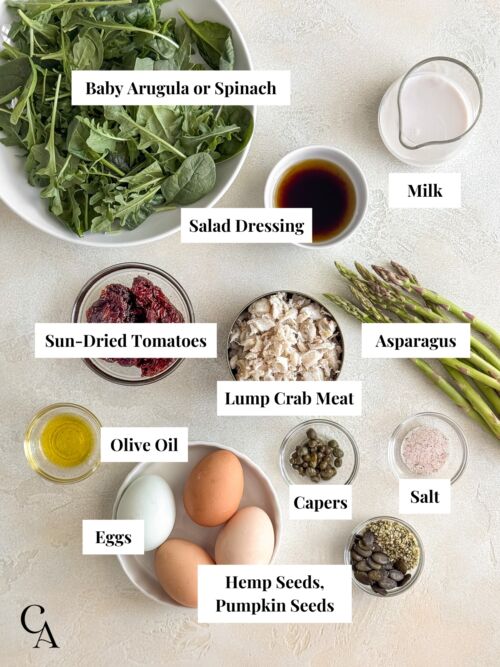Bowls of ingredients on a light surface, including lump crab meat, eggs, asparagus, sun-dried tomatoes, and baby greens.