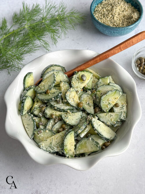 A shallow, white bowl of cucumber salad with dill and garlic aioli sauce, with bowls of capers and hemp seeds in the background.